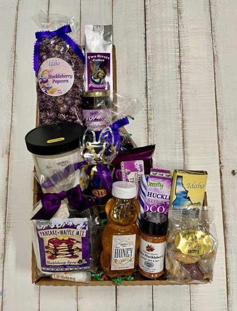 IMG_4151+(1) A beautifully arranged gift basket featuring Idaho huckleberry products, including huckleberry popcorn, pancake and waffle mix, huckleberry syrup, honey, and assorted snacks. The display is adorned with purple accents and includes coffee and chocolate items, making it an ideal gift for food lovers and huckleberry enthusiasts.