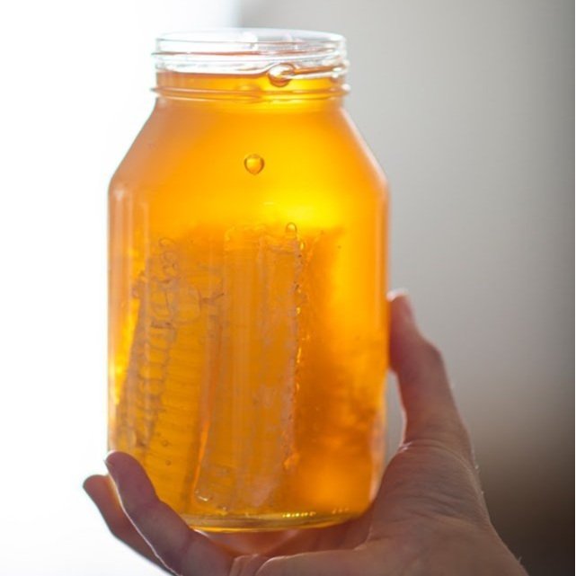 Hand holding a clear jar filled with golden honey and honeycomb, illuminated by natural light, showcasing the rich texture and color of the honey.