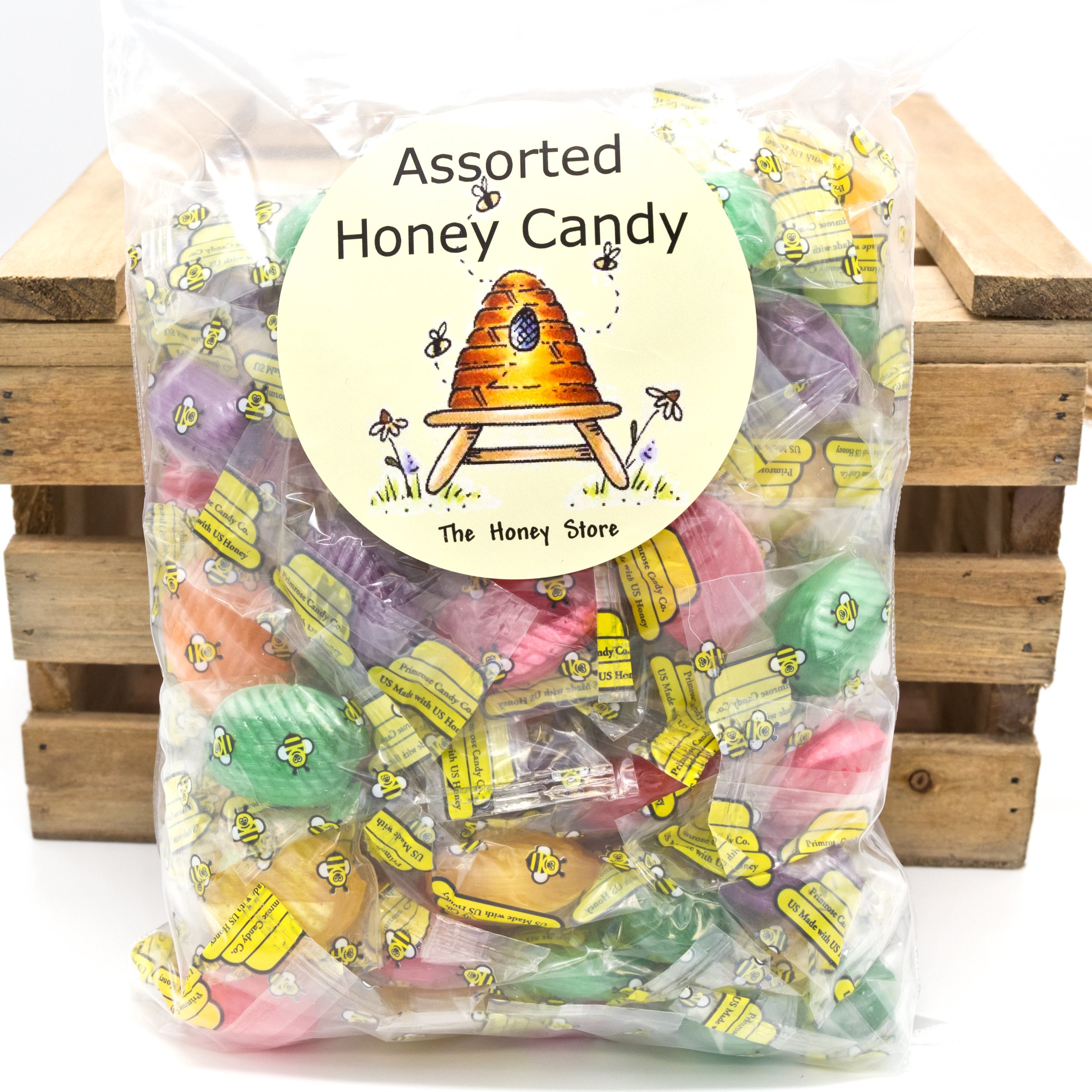 The+Honey+Store+-+HoneyCandy_assorted_1745 Clear plastic bag filled with assorted honey candy, featuring vibrant colors and a label with a bee and a beehive illustration, resting against a wooden crate.