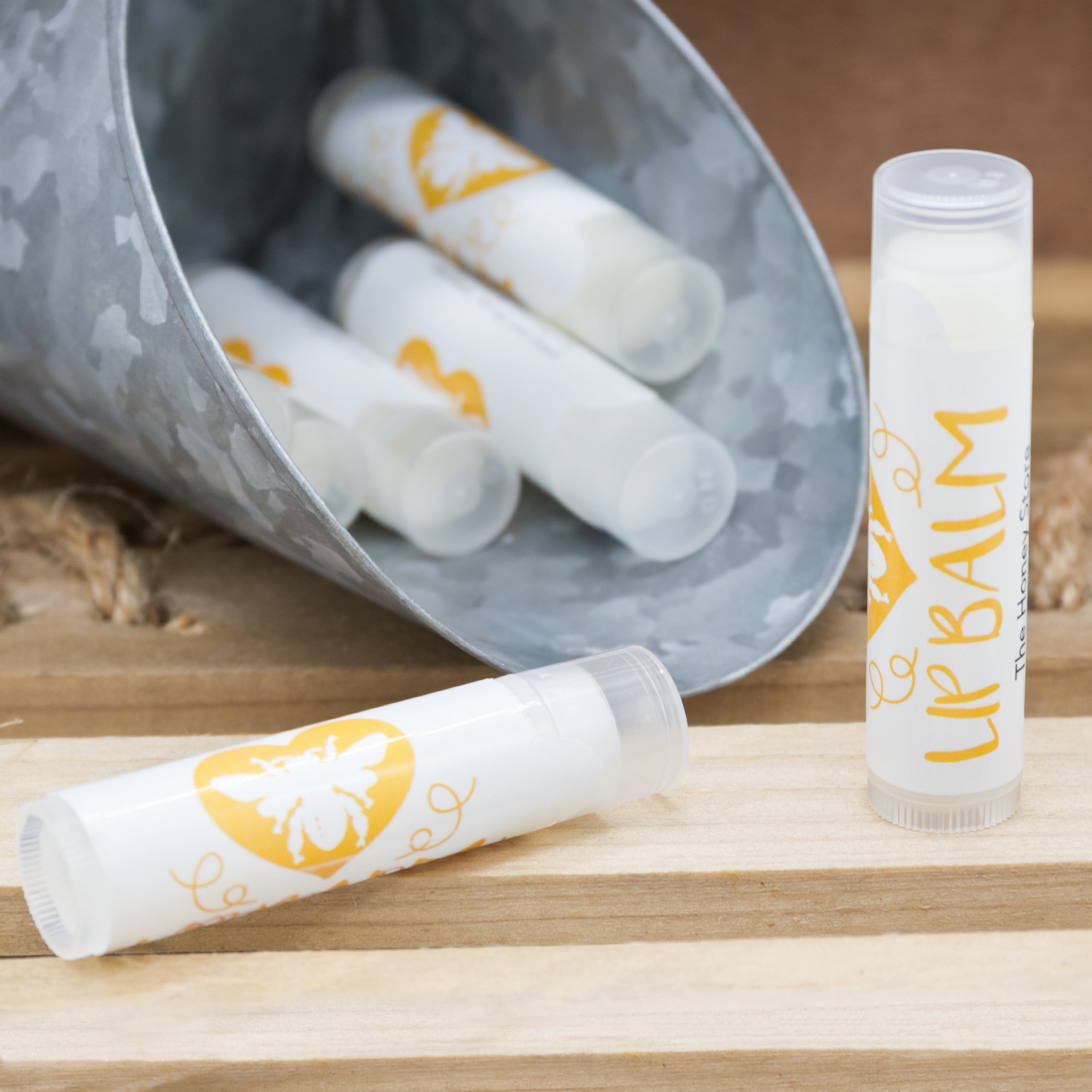 The+Honey+Store+-+LipBalm_honey_display_1770 Lip balm tubes with a bee logo are displayed, some spilling out of a metal container onto a wooden surface, showcasing a natural and eco-friendly product.