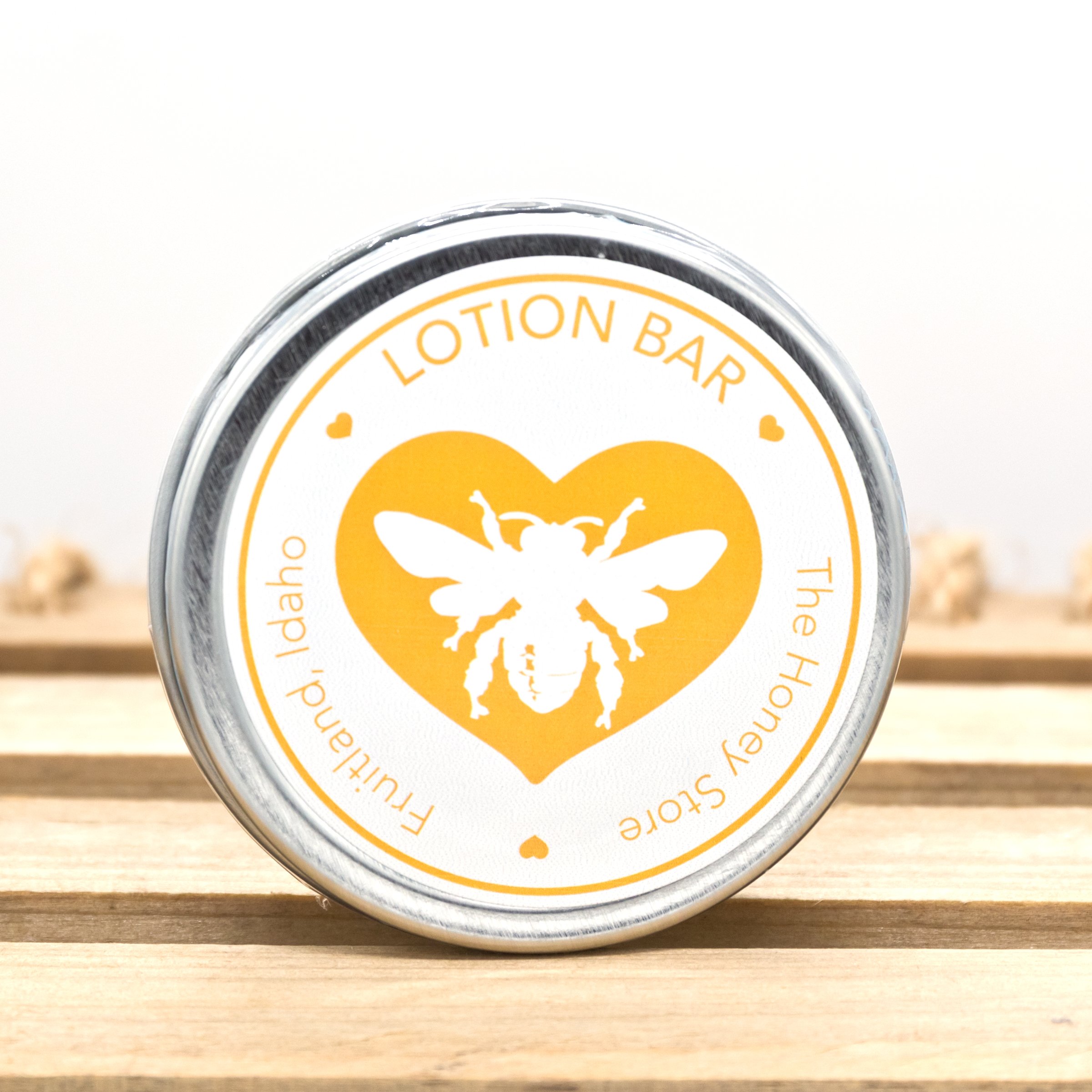 Lotion bar in a metal tin with a bee logo and heart design, labeled "The Honey Store, Fruitland, Idaho," showcasing natural skincare product for moisturizing.